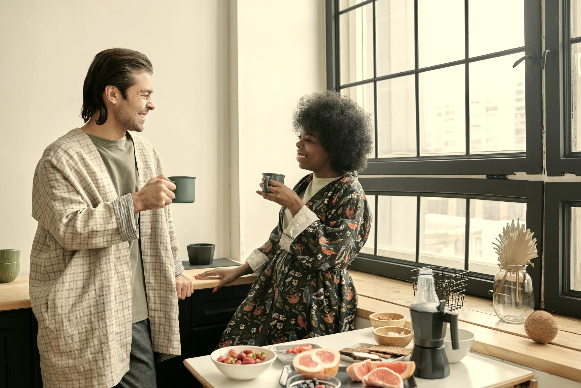 Two people at a kitchen table in morning light, coffee between them